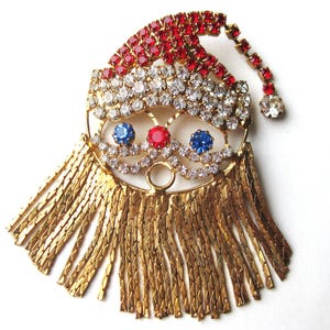 May include: A gold-tone metal brooch shaped like a Santa Claus face. The hat is encrusted with red and clear rhinestones. The eyes are blue rhinestones, the nose is a red rhinestone, and the beard consists of long, dangling gold-tone metal chains.
