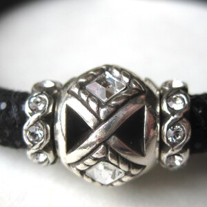 Puede incluir: Una pulsera negra texturizada con una cuenta central plateada con dise&ntilde;o de cruz, incrustada con cristales cuadrados transparentes. La cuenta central est&aacute; flanqueada por dos cuentas espaciadoras plateadas, cada una con varios cristales redondos.