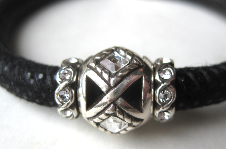 Puede incluir: Una pulsera negra texturizada con una cuenta central plateada con dise&ntilde;o de cruz, incrustada con cristales cuadrados transparentes. La cuenta central est&aacute; flanqueada por dos cuentas espaciadoras plateadas, cada una con varios cristales redondos.