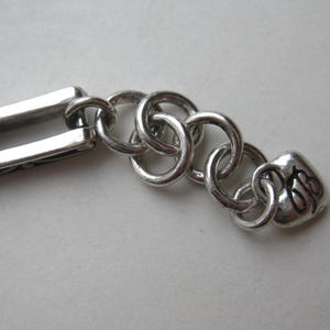 May include: A silver-toned metal bracelet extension chain featuring a series of interlocking circular links connected to a rectangular link on one end and a small heart-shaped charm with the black engraved letters "JB" on the other end.