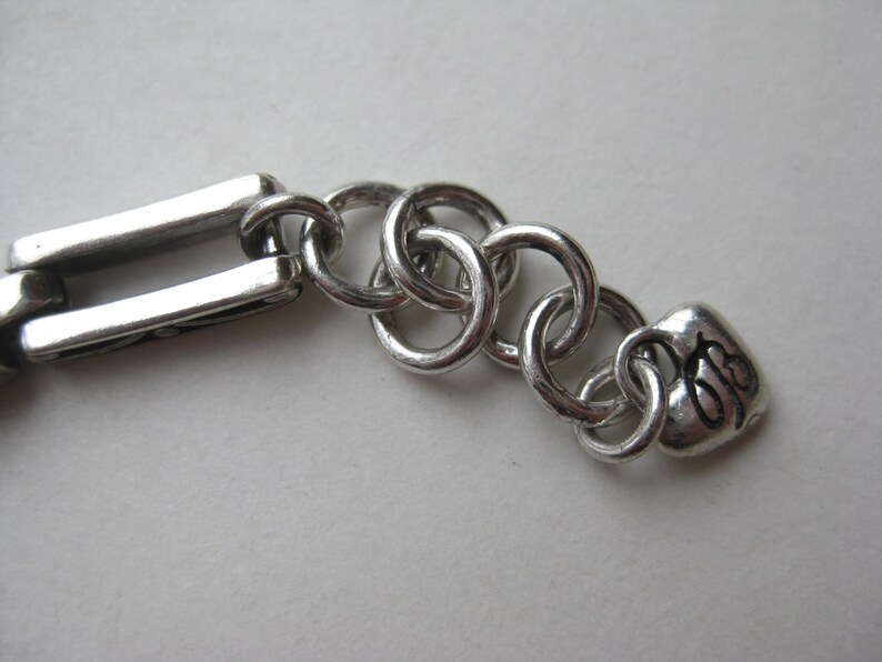 May include: A silver-toned metal bracelet extension chain featuring a series of interlocking circular links connected to a rectangular link on one end and a small heart-shaped charm with the black engraved letters "JB" on the other end.