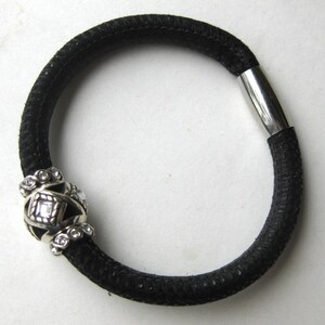 Puede incluir: Una pulsera de cord&oacute;n negro texturizado con un cierre de metal plateado y una cuenta central decorativa plateada. La cuenta tiene un dise&ntilde;o geom&eacute;trico de jaula con cristales transparentes incrustados, contrastando con la banda oscura y flexible.