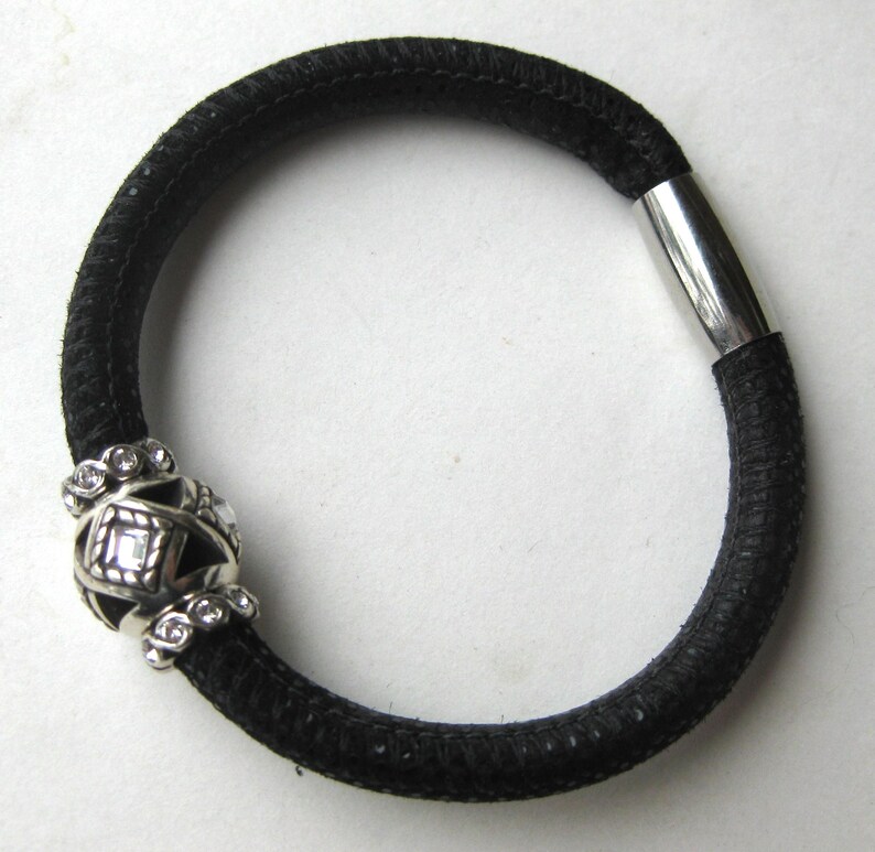 Puede incluir: Una pulsera de cord&oacute;n negro texturizado con un cierre de metal plateado y una cuenta central decorativa plateada. La cuenta tiene un dise&ntilde;o geom&eacute;trico de jaula con cristales transparentes incrustados, contrastando con la banda oscura y flexible.