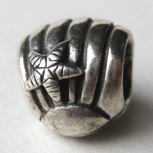 May include: A silver charm featuring a starfish design on a ribbed, rounded shape. The starfish is detailed with etched lines, and the charm has a hole for stringing. The charm is likely made of sterling silver and is a beach-themed accessory.