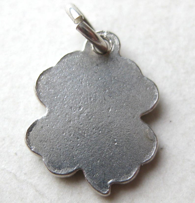 K&ouml;nnte beinhalten: Ein silberfarbener Kleeblatt-Anh&auml;nger mit strukturierter Oberfl&auml;che. Der Anh&auml;nger hat einen gewellten Rand und eine kleine Schlaufe oben zum Befestigen an einer Halskette oder einem Armband. Der Anh&auml;nger ist ein vierbl&auml;ttriges Kleeblatt.