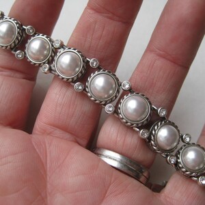Puede incluir: Una pulsera de tono plateado que presenta una serie de perlas blancas redondas engastadas en bordes de metal trenzado. Cada engaste de perla est&aacute; conectado por peque&ntilde;os diamantes de imitaci&oacute;n claros y brillantes en un patr&oacute;n.