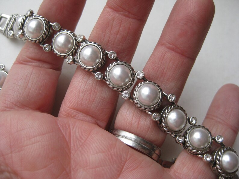 Puede incluir: Una pulsera de tono plateado que presenta una serie de perlas blancas redondas engastadas en bordes de metal trenzado. Cada engaste de perla est&aacute; conectado por peque&ntilde;os diamantes de imitaci&oacute;n claros y brillantes en un patr&oacute;n.