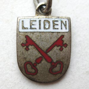 May include: A shield-shaped metal charm featuring a white enamel band at the top with the word LEIDEN in black letters. Below the text, two red enamel keys are crossed against a textured silver-toned background. A metal jump ring is attached.
