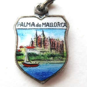 May include: A silver-toned shield-shaped pendant with a painted scene of Palma de Mallorca. The image features a red sailboat on blue water, a detailed building, and the text "PALMA de MALLORCA" at the top. The pendant has a silver loop for attachment.