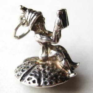 May include: A silver charm depicting a seated gnome holding a mug, wearing a pointed hat, and seated on a mushroom-shaped base. The charm is detailed with etched lines and dots, suggesting a vintage or antique style.