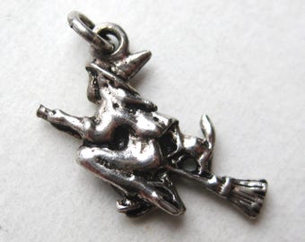May include: A silver-toned metal charm depicting a figure wearing a pointed hat and riding a broomstick, accompanied by a small animal figure positioned behind the rider. The charm includes a circular jump ring at the top for attachment.