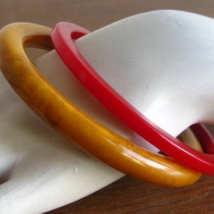 Può includere: Due braccialetti rigidi rotondi, uno di colore rosso brillante solido e l'altro giallo ambra traslucido con una consistenza marmorizzata e vorticosa. Entrambi i braccialetti presentano una finitura liscia e lucida e un design circolare semplice.