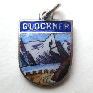 以下が含まれることがあります： 白でGLOCKNERと書かれた青いエナメルのヘッダーが付いた、盾形の銀色の金属製チャーム。中央には、白い山頂、紫色の岩層、茶色の前景と柵が描かれた山岳風景がペイントされています。