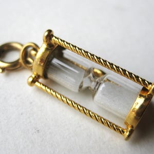 May include: A gold-colored hourglass charm with a clear glass center filled with white sand. The hourglass is framed by twisted gold bars and has a small clasp for attaching to a necklace or bracelet. The charm is a miniature representation of time.