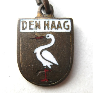 May include: A bronze-toned metal charm shaped like a shield, featuring a white enamel stork holding a small red and black object in its beak. The top section has a white enamel banner with the black text DEN HAAG. A metal loop is attached at the top.