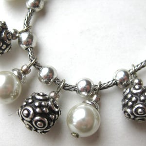 Puede incluir: Pulsera plateada con cadena trenzada y abalorios colgantes. Los abalorios incluyen cuentas redondas plateadas, cuentas plateadas texturizadas y perlas de imitaci&oacute;n blancas nacaradas. Primer plano sobre fondo blanco.