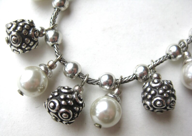 Puede incluir: Pulsera plateada con cadena trenzada y abalorios colgantes. Los abalorios incluyen cuentas redondas plateadas, cuentas plateadas texturizadas y perlas de imitaci&oacute;n blancas nacaradas. Primer plano sobre fondo blanco.