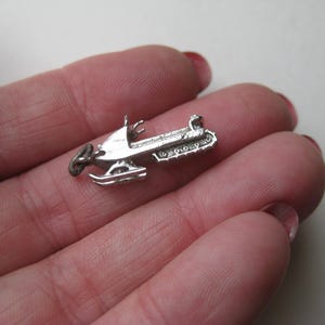 Może przedstawiać: Mały, srebrny wisiorek w kształcie skutera śnieżnego. Urok jest szczeg&oacute;łowy z nartami, siedzeniem i gąsienicą. Skuter śnieżny jest pokazany na neutralnym tle. Ten urok może być używany do tworzenia biżuterii lub jako przedmiot kolekcjonerski.