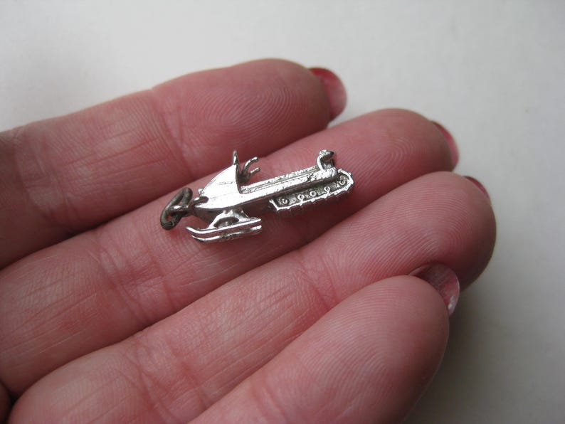 Może przedstawiać: Mały, srebrny wisiorek w kształcie skutera śnieżnego. Urok jest szczeg&oacute;łowy z nartami, siedzeniem i gąsienicą. Skuter śnieżny jest pokazany na neutralnym tle. Ten urok może być używany do tworzenia biżuterii lub jako przedmiot kolekcjonerski.