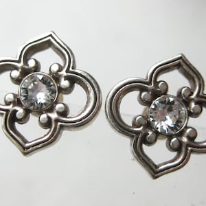 May include: A pair of silver-toned metal stud earrings featuring a four-lobed openwork design. Each earring has a circular clear faceted crystal set in the center, surrounded by a decorative frame with rounded edges and symmetrical cutouts.