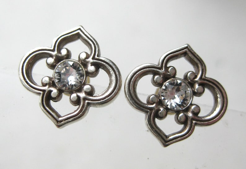 May include: A pair of silver-toned metal stud earrings featuring a four-lobed openwork design. Each earring has a circular clear faceted crystal set in the center, surrounded by a decorative frame with rounded edges and symmetrical cutouts.