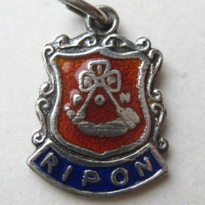 Pode incluir: Pingente prateado com design de escudo. O escudo apresenta um fundo de esmalte vermelho com um emblema branco e a palavra "RIPON" em esmalte azul. O pingente tem uma borda ornamentada e uma pequena argola para fixação.