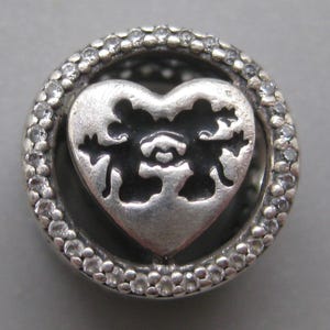 May include: A circular silver charm featuring a central heart cutout with a silhouette of two mice facing each other. The outer ring is encrusted with small, clear, round stones, creating a textured border around the central heart design.