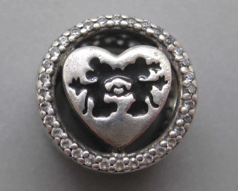 May include: A circular silver charm featuring a central heart cutout with a silhouette of two mice facing each other. The outer ring is encrusted with small, clear, round stones, creating a textured border around the central heart design.