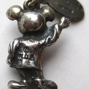 May include: A silver-toned charm in the shape of a cartoon character with a round head, arms outstretched, and wearing a shirt and pants. The charm has a loop at the top for attachment and a small tag. The character's face is not visible.
