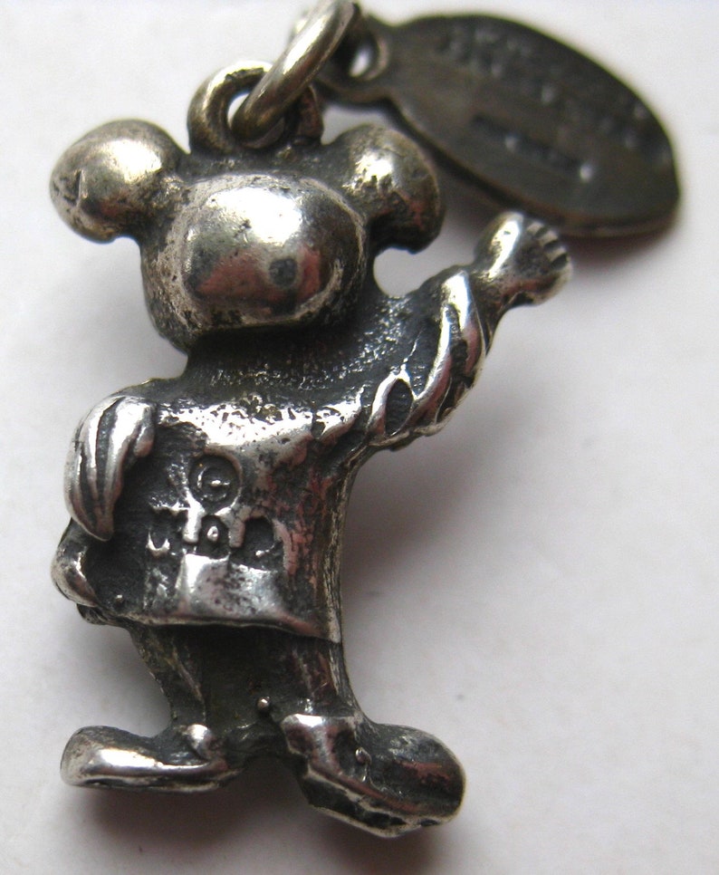 May include: A silver-toned charm in the shape of a cartoon character with a round head, arms outstretched, and wearing a shirt and pants. The charm has a loop at the top for attachment and a small tag. The character's face is not visible.