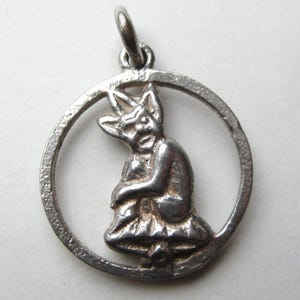 May include: A silver pendant featuring a seated devil-like figure within a circular frame. The figure has pointed ears and a stern expression. The pendant is detailed with visible textures and a loop for a chain.