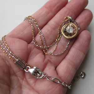 Puede incluir: Un collar de m&uacute;ltiples hilos con una mezcla de cadenas met&aacute;licas en tonos oro y plata. El collar incluye un colgante con una piedra tallada en forma de coj&iacute;n de color marr&oacute;n claro, engastada en un bisel dorado y un cierre de mosquet&oacute;n plateado.