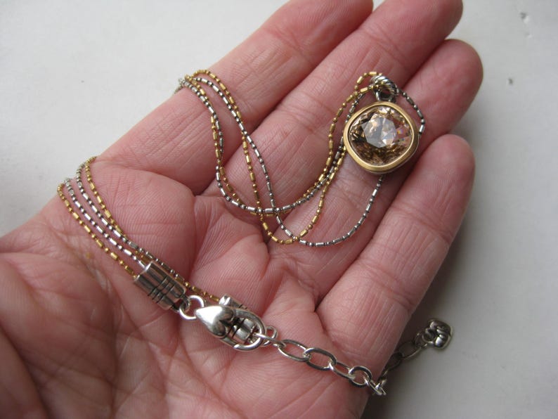 Puede incluir: Un collar de m&uacute;ltiples hilos con una mezcla de cadenas met&aacute;licas en tonos oro y plata. El collar incluye un colgante con una piedra tallada en forma de coj&iacute;n de color marr&oacute;n claro, engastada en un bisel dorado y un cierre de mosquet&oacute;n plateado.