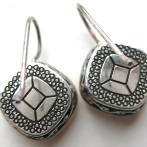 May include: A pair of silver-toned drop earrings featuring a square-shaped design with a central square motif surrounded by a dotted border. The sides of the earrings display an intricate scrollwork pattern, and each piece is attached to a curved wire hook.