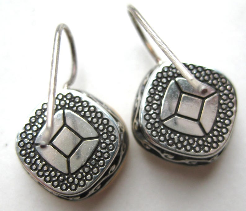 May include: A pair of silver-toned drop earrings featuring a square-shaped design with a central square motif surrounded by a dotted border. The sides of the earrings display an intricate scrollwork pattern, and each piece is attached to a curved wire hook.
