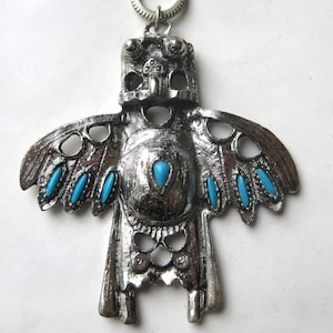 May include: A silver-toned metal pendant shaped like a stylized bird with outstretched wings. The pendant features a textured surface and is inlaid with small, oval-shaped bright blue stones along the wings and a single teardrop stone at the center.