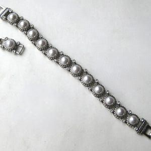 Puede incluir: Una pulsera de eslabones de metal plateado con una serie de perlas blancas redondas engastadas en marcos circulares texturizados con estilo de cuerda. La pulsera incluye peque&ntilde;as piedras transparentes entre cada perla y un cierre de metal seguro.