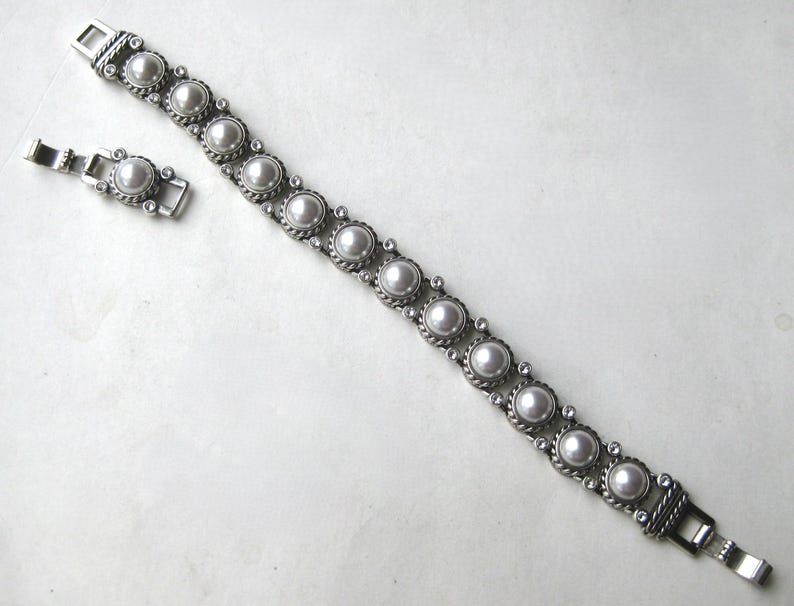 Puede incluir: Una pulsera de eslabones de metal plateado con una serie de perlas blancas redondas engastadas en marcos circulares texturizados con estilo de cuerda. La pulsera incluye peque&ntilde;as piedras transparentes entre cada perla y un cierre de metal seguro.