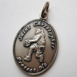 Op de afbeelding: Zilveren ovale hanger met een reliëf van Sint-Christoffel, die een basketbal vasthoudt. De woorden "Saint Christopher Protect Me" zijn rond de rand gegraveerd. De hanger heeft een kleine lus aan de bovenkant voor een ketting.