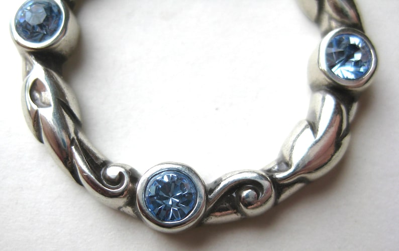 May include: A silver-toned ring with a braided design, featuring four round, light blue gemstones. The gemstones are set in individual bezels, and the ring has a polished finish. The ring is a piece of jewelry.