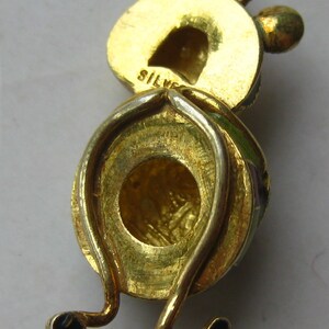 May include: A gold-toned metal charm shaped like a cartoon character with a round head, two small ears, and thin legs ending in black feet. The word SILVER is stamped on the back of the head. The charm features a circular loop at the top.