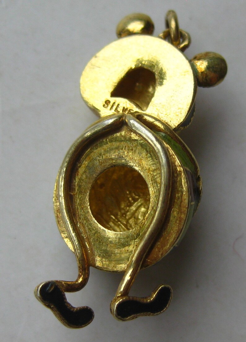 May include: A gold-toned metal charm shaped like a cartoon character with a round head, two small ears, and thin legs ending in black feet. The word SILVER is stamped on the back of the head. The charm features a circular loop at the top.