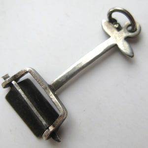 May include: A silver-toned charm in the shape of a mandolin. The charm has a rectangular body with a dark, possibly black, insert, and a long handle ending in a decorative loop. The charm is likely made of metal.