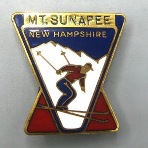 May include: A trapezoidal gold-tone metal pin featuring a skier in a red top and blue pants on a white mountain background. The pin has blue and red enamel accents and includes the text MT. SUNAPEE and NEW HAMPSHIRE in gold lettering.