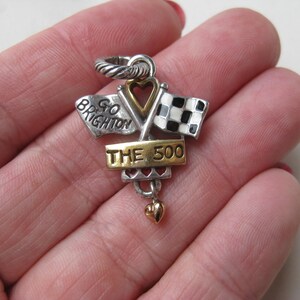 May include: Silver and gold-toned charm with a racing theme. Features two crossed flags, one checkered and one with text "GO BRIGHTON". The charm includes the text "THE 500" and a small heart pendant.
