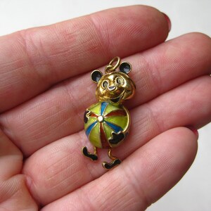 May include: A gold-toned metal pendant shaped like a cartoon bear holding a multi-colored ball. The bear features black enamel accents on its ears, hands, and feet, while the ball is decorated with segments of red, blue, and green enamel.