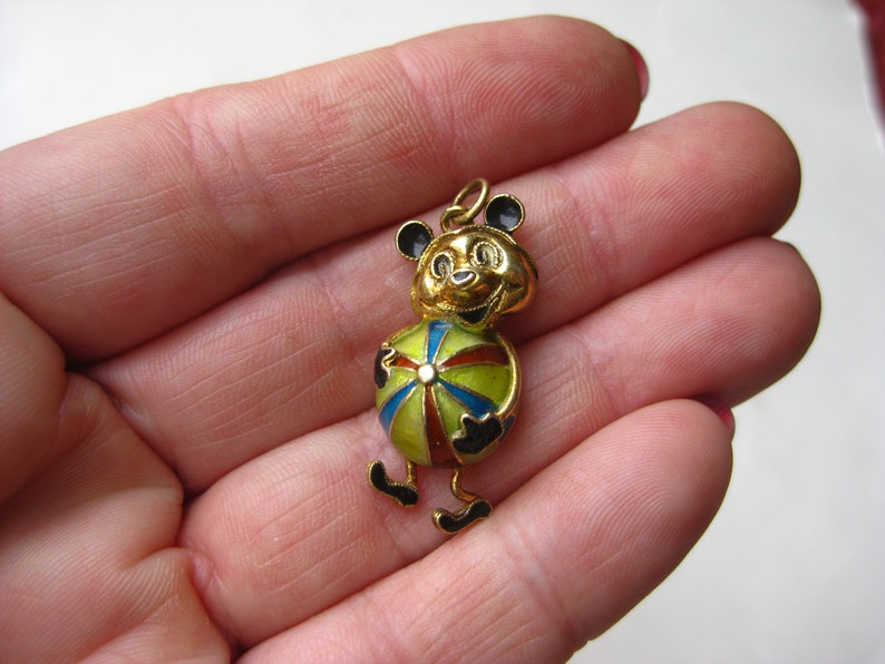 May include: A gold-toned metal pendant shaped like a cartoon bear holding a multi-colored ball. The bear features black enamel accents on its ears, hands, and feet, while the ball is decorated with segments of red, blue, and green enamel.