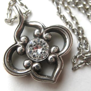 May include: A silver-toned metal pendant necklace featuring a four-lobed, clover-like frame with rounded edges. A single, clear, round faceted gemstone is set in the center of the pendant, which hangs from a matching silver-toned link chain.