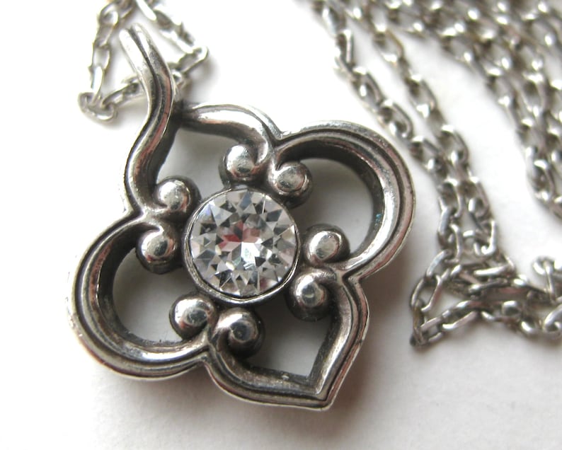 May include: A silver-toned metal pendant necklace featuring a four-lobed, clover-like frame with rounded edges. A single, clear, round faceted gemstone is set in the center of the pendant, which hangs from a matching silver-toned link chain.
