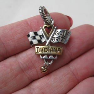 May include: A silver and gold charm with the word "INDIANA" on a gold rectangular plate. The charm features crossed racing flags, a heart-shaped cutout, and the words "GO BRIGHTON". A small gold heart dangles below.
